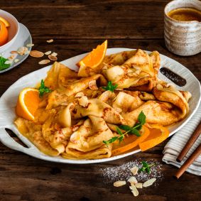 Crêpes Suzette auf einer Servierplatte, garniert mit Orangenscheiben, Mandelblättchen und Minze, serviert mit Orangensauce und zwei Tassen Kaffee auf einem rustikalen Holztisch.