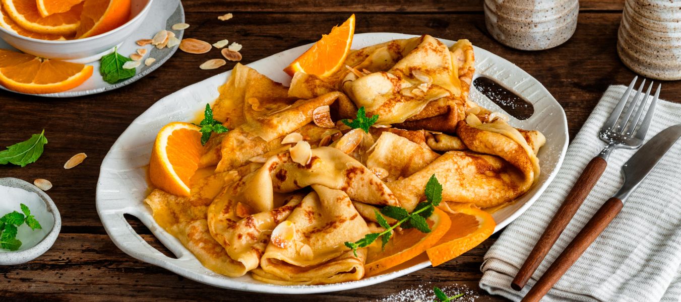 Crêpes Suzette auf einer Servierplatte, garniert mit Orangenscheiben, Mandelblättchen und Minze, serviert mit Orangensauce und zwei Tassen Kaffee auf einem rustikalen Holztisch.