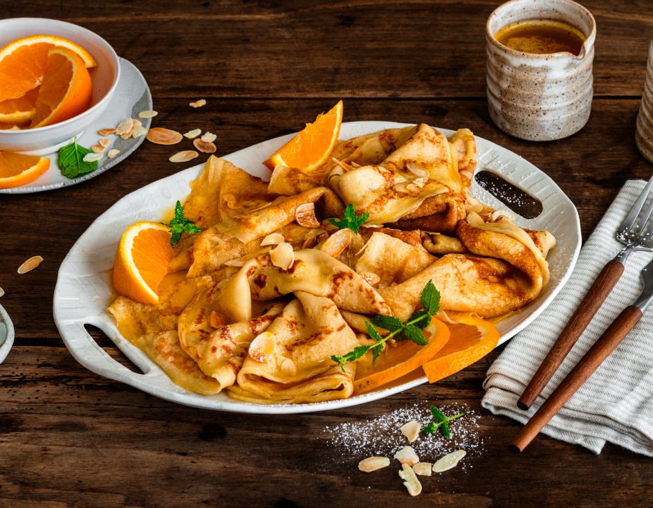 Crêpes Suzette auf einer Servierplatte, garniert mit Orangenscheiben, Mandelblättchen und Minze, serviert mit Orangensauce und zwei Tassen Kaffee auf einem rustikalen Holztisch.