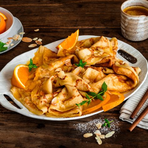 Crêpes Suzette auf einer Servierplatte, garniert mit Orangenscheiben, Mandelblättchen und Minze, serviert mit Orangensauce und zwei Tassen Kaffee auf einem rustikalen Holztisch.