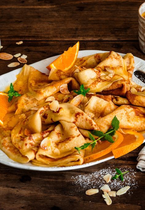Crêpes Suzette auf einer Servierplatte, garniert mit Orangenscheiben, Mandelblättchen und Minze, serviert mit Orangensauce und zwei Tassen Kaffee auf einem rustikalen Holztisch.