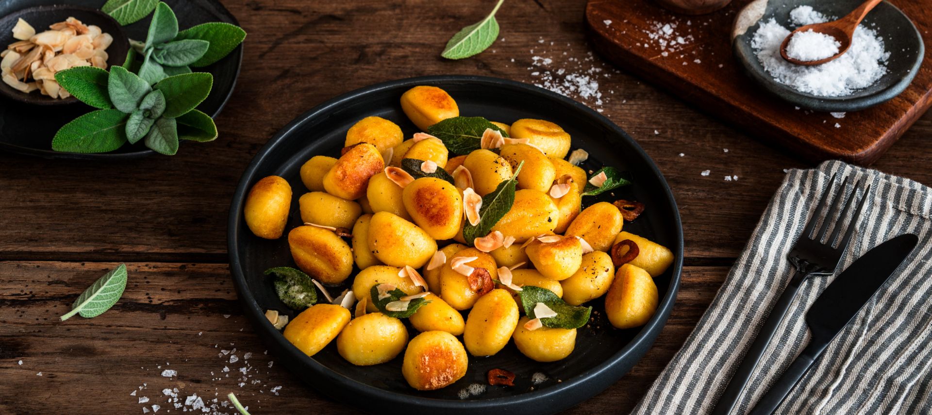 Gebratene Gnocchi mit Salbeiblättern und gerösteten Mandelblättchen auf einem schwarzen Teller auf rustikalem Holztisch, umgeben von Salz, frischem Salbei sowie Besteck auf einer Stoffserviette.