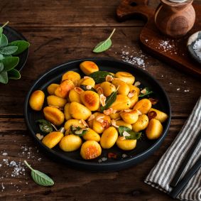 Gebratene Gnocchi mit Salbeiblättern und gerösteten Mandelblättchen auf einem schwarzen Teller auf rustikalem Holztisch, umgeben von Salz, frischem Salbei sowie Besteck auf einer Stoffserviette.