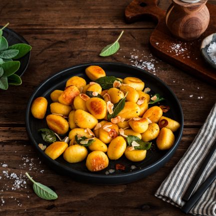 Gebratene Gnocchi mit Salbeiblättern und gerösteten Mandelblättchen auf einem schwarzen Teller auf rustikalem Holztisch, umgeben von Salz, frischem Salbei sowie Besteck auf einer Stoffserviette.