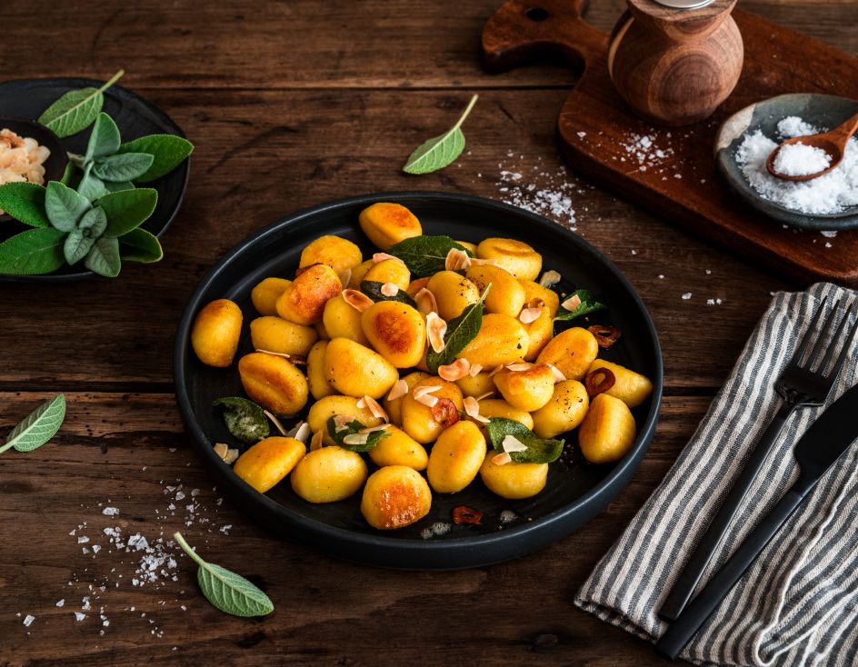 Gebratene Gnocchi mit Salbeiblättern und gerösteten Mandelblättchen auf einem schwarzen Teller auf rustikalem Holztisch, umgeben von Salz, frischem Salbei sowie Besteck auf einer Stoffserviette.