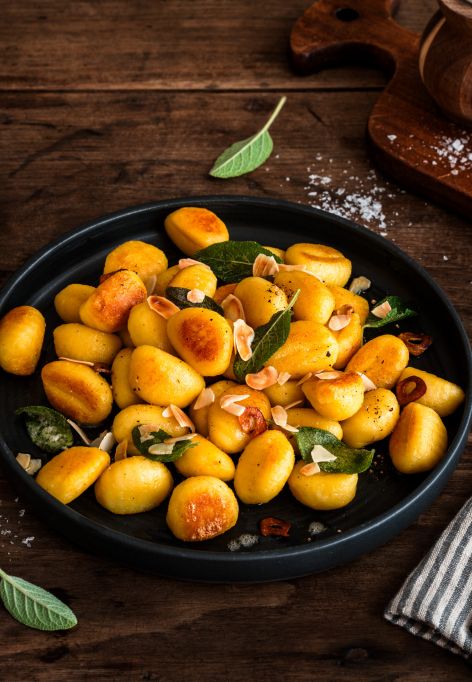 Gebratene Gnocchi mit Salbeiblättern und gerösteten Mandelblättchen auf einem schwarzen Teller auf rustikalem Holztisch, umgeben von Salz, frischem Salbei sowie Besteck auf einer Stoffserviette.
