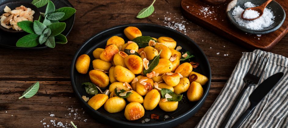 Gebratene Gnocchi mit Salbeiblättern und gerösteten Mandelblättchen auf einem schwarzen Teller auf rustikalem Holztisch, umgeben von Salz, frischem Salbei sowie Besteck auf einer Stoffserviette.