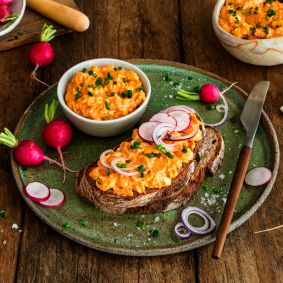 Rustikales Bauernbrot mit cremigem Obatzda, garniert mit Radieschenscheiben, roten Zwiebelringen und Schnittlauch auf einem grünen Teller. Daneben stehen eine Schale mit Obatzda sowie frische Radieschen auf einem Holztisch.