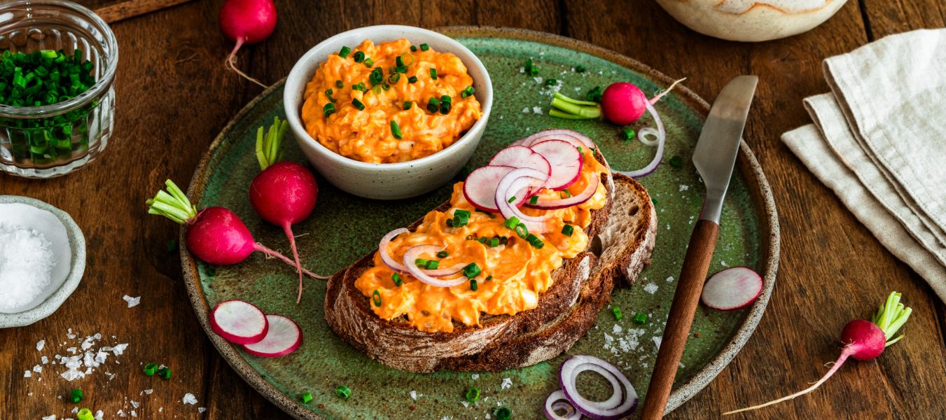 Rustikales Bauernbrot mit cremigem Obatzda, garniert mit Radieschenscheiben, roten Zwiebelringen und Schnittlauch auf einem grünen Teller. Daneben stehen eine Schale mit Obatzda sowie frische Radieschen auf einem Holztisch.