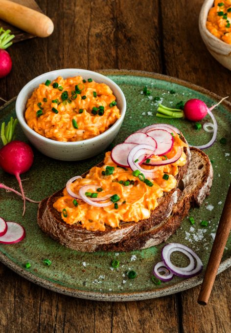 Rustikales Bauernbrot mit cremigem Obatzda, garniert mit Radieschenscheiben, roten Zwiebelringen und Schnittlauch auf einem grünen Teller. Daneben stehen eine Schale mit Obatzda sowie frische Radieschen auf einem Holztisch.