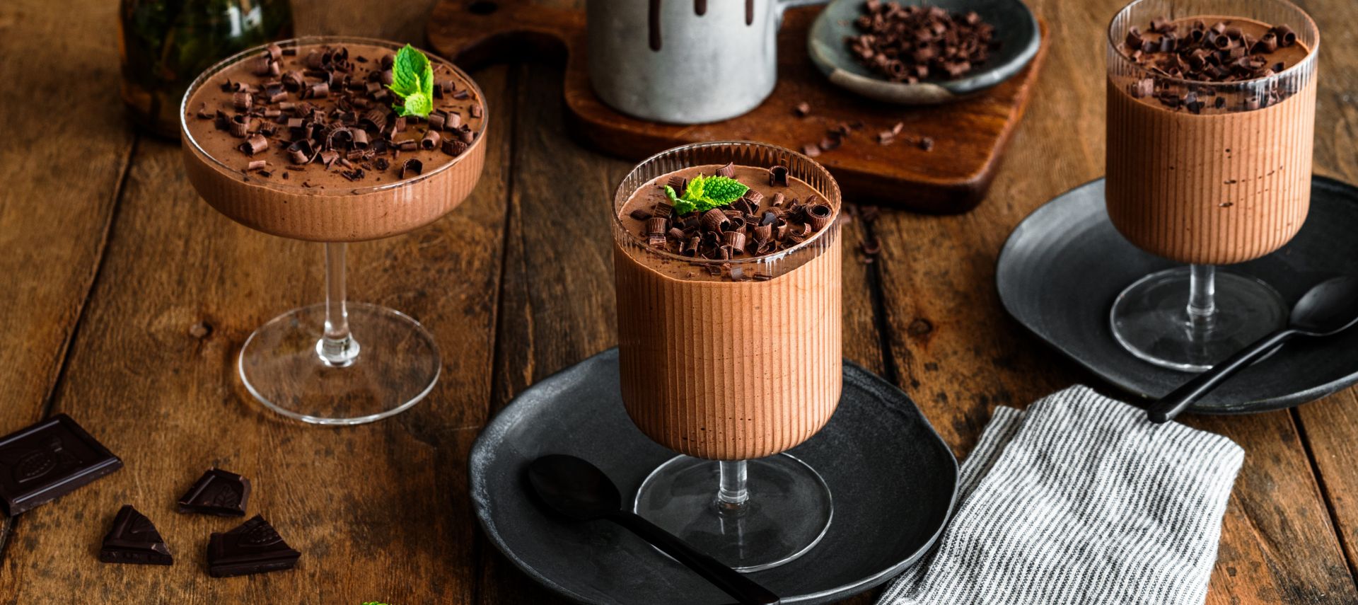 Zwei Gläser mit cremigem Schokoladenmousse, garniert mit dunklen Schokoladenraspeln und einem frischen Minzblatt.