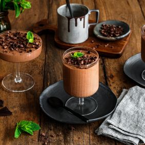 Zwei Gläser mit cremigem Schokoladenmousse, garniert mit dunklen Schokoladenraspeln und einem frischen Minzblatt.