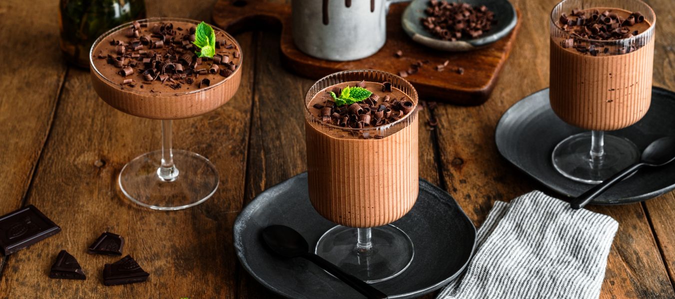 Zwei Gläser mit cremigem Schokoladenmousse, garniert mit dunklen Schokoladenraspeln und einem frischen Minzblatt.