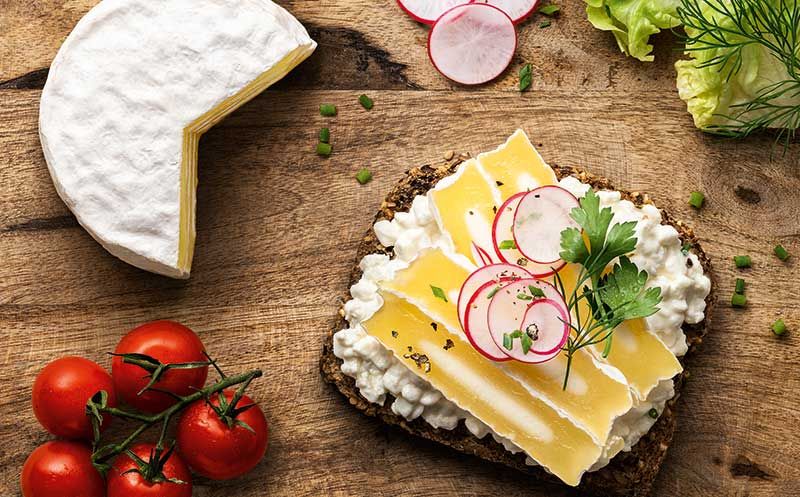 Rustikales Holzbrett mit einem belegten Vollkornbrot, garniert mit Hüttenkäse, Käsescheiben, Radieschen und frischen Kräutern.