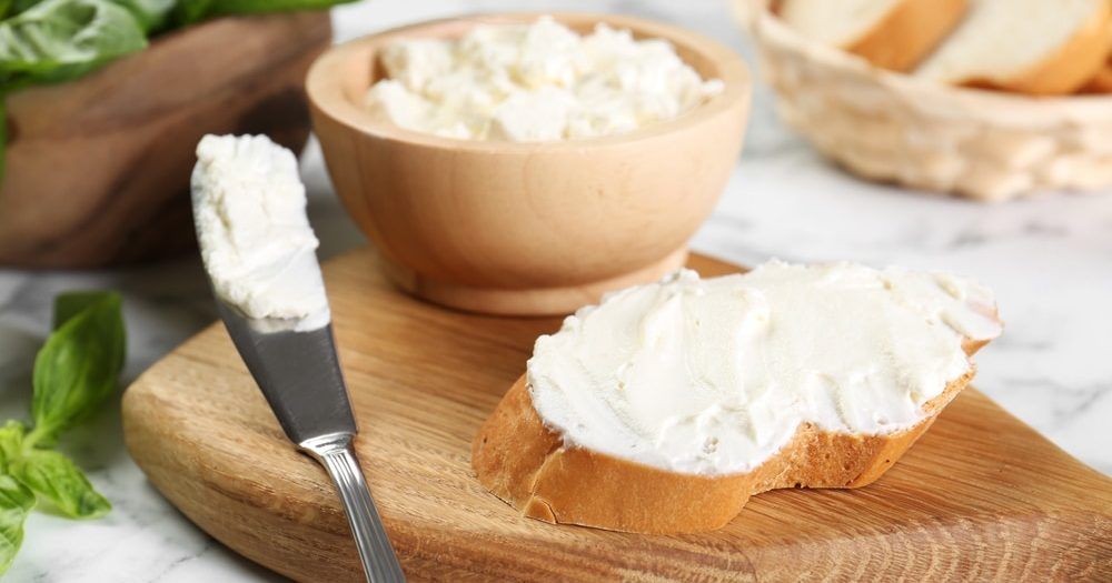 Frisches Weißbrot mit cremigem Frischkäse bestrichen, daneben ein Messer mit Käse und eine Holzschale mit weiterer Portion Frischkäse.
