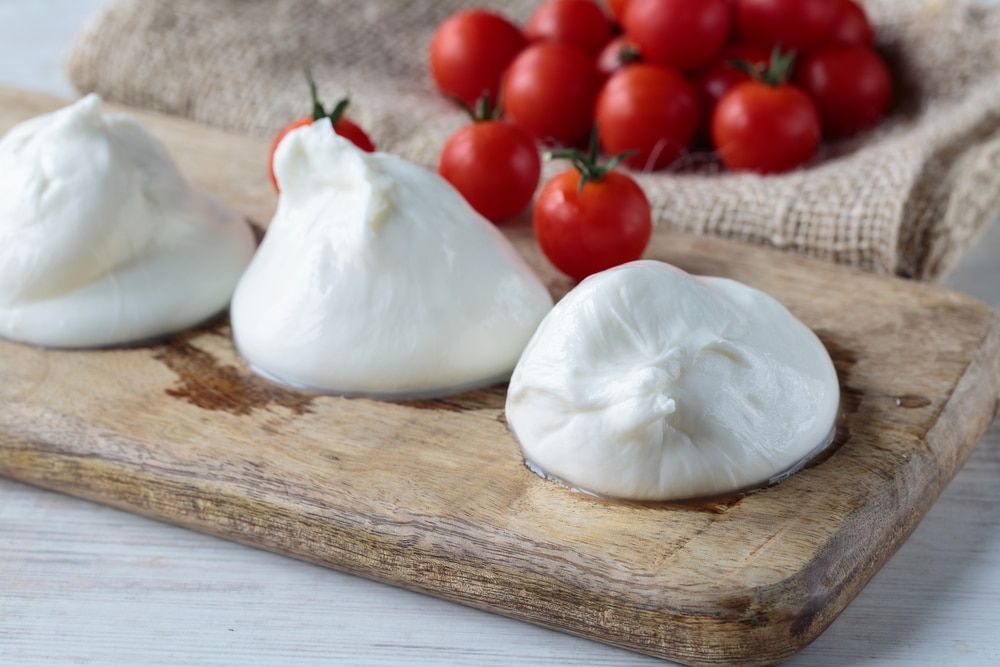 Drei frische Burrata-Käsekugeln auf einem Holzbrett, im Hintergrund leuchtend rote Kirschtomaten auf einem Jutesack.