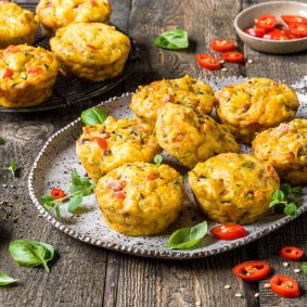 Mini-Gemüsefrittatas, die an herzhafte Muffins erinnern, werden auf einem rustikalen Teller serviert, garniert mit frischen Kräutern und geschnittenen roten Chilischoten. Weitere Frittatas und Schüsseln mit Gewürzen stehen im Hintergrund auf einem Holztisch.