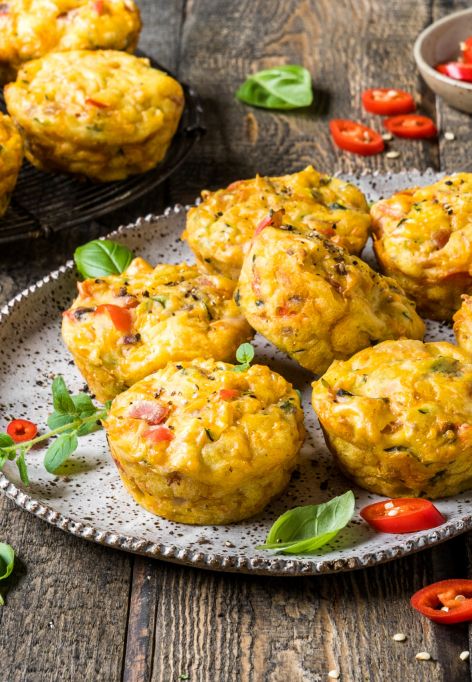 Mini-Gemüsefrittatas, die an herzhafte Muffins erinnern, werden auf einem rustikalen Teller serviert, garniert mit frischen Kräutern und geschnittenen roten Chilischoten. Weitere Frittatas und Schüsseln mit Gewürzen stehen im Hintergrund auf einem Holztisch.