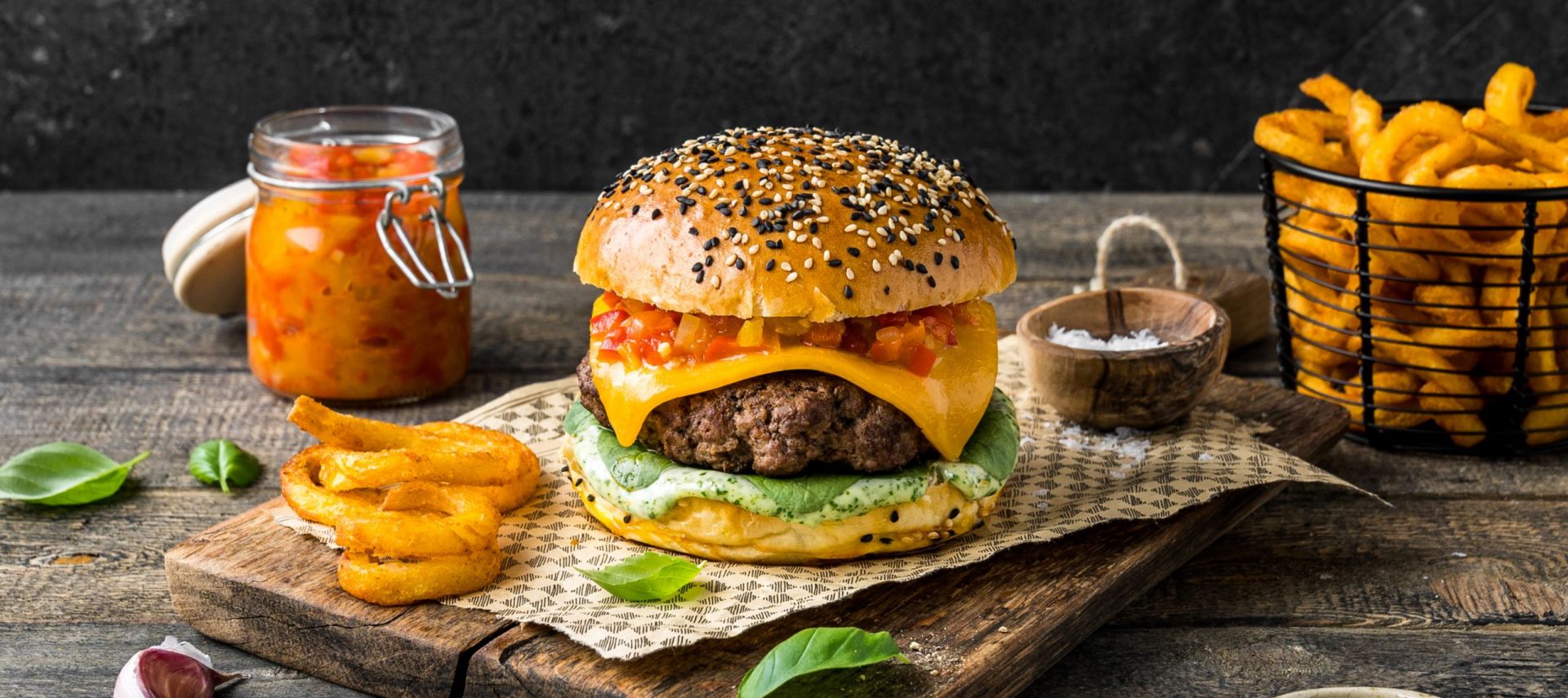 Ein Cheeseburger mit Salat, Cheddar-Käse und Relish auf einem Brötchen mit Körnern liegt auf einem Holzbrett, neben Curly Fries, einer kleinen Schüssel Salz, einem Glas Relish und einem Korb mit Curly Fries.