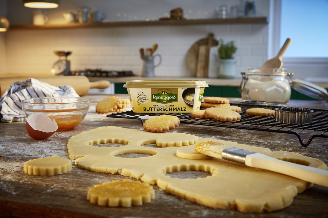 Eine Küchenszene mit Keksteig, der auf einer bemehlten Oberfläche geschnitten wird, einem Becher Kerrygold-Butterschmalz, einem geschnittenen Ei, einer Schüssel Glasur, einem Backpinsel, abkühlenden Keksen auf einem Rost und Backutensilien im Hintergrund.
