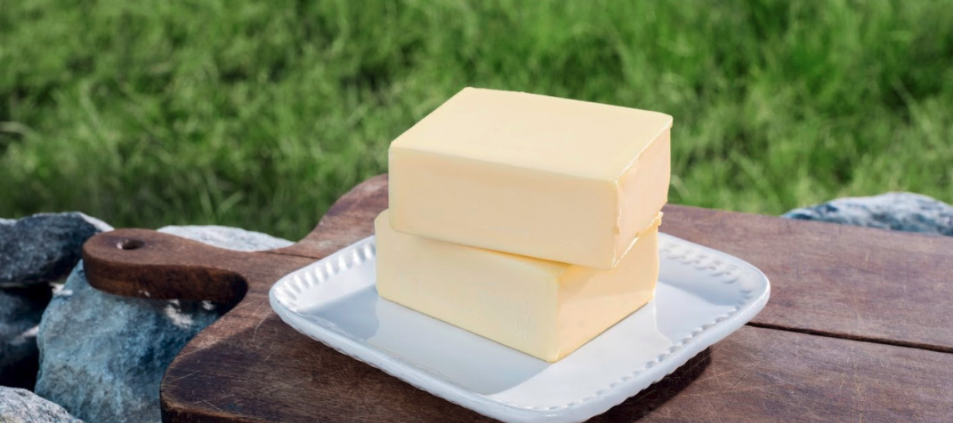 Zwei Butterstücke auf einem weißen Teller gestapelt, auf einem hölzernen Schneidebrett im Freien mit grünem Gras und Felsen im Hintergrund platziert.