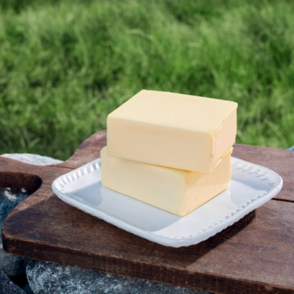 Zwei Butterstücke auf einem weißen Teller gestapelt, auf einem hölzernen Schneidebrett im Freien mit grünem Gras und Felsen im Hintergrund platziert.