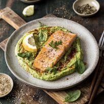 Gegrilltes Lachsfilet auf grünem Hummus, garniert mit einer Zitronenscheibe, frischem Spinat, Petersilie, Sesamsamen und schwarzem Pfeffer auf einem Keramikteller. Zitronenscheiben, Gewürze und Besteck liegen bereit.