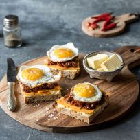Drei belegte Brötchen mit geschmolzenem Käse, karamellisierten Zwiebeln und Spiegeleiern auf einem Holzbrett, daneben eine Schüssel Streichfette, ein Messer und Gewürze. Im Hintergrund rote Chilischoten und Gewürze.