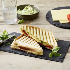Ein halbiertes Panini mit gegrilltem Käse und Tomaten liegt mit frischem Basilikum auf einer Schieferplatte. Im Hintergrund stehen Cheddar-Sandwiches, rote Paprika, eine Schüssel Brotaufstrich, zwei Gläser Wasser und eine kleine Schüssel Salz.