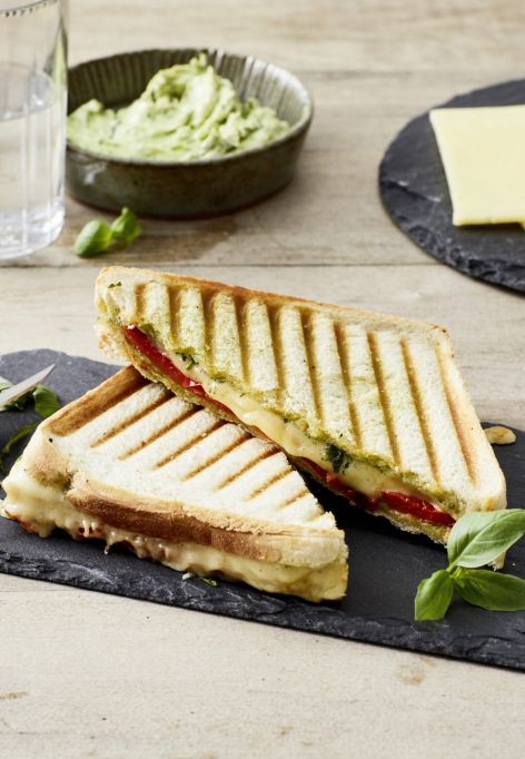 Ein halbiertes Panini mit gegrilltem Käse und Tomaten liegt mit frischem Basilikum auf einer Schieferplatte. Im Hintergrund stehen Cheddar-Sandwiches, rote Paprika, eine Schüssel Brotaufstrich, zwei Gläser Wasser und eine kleine Schüssel Salz.