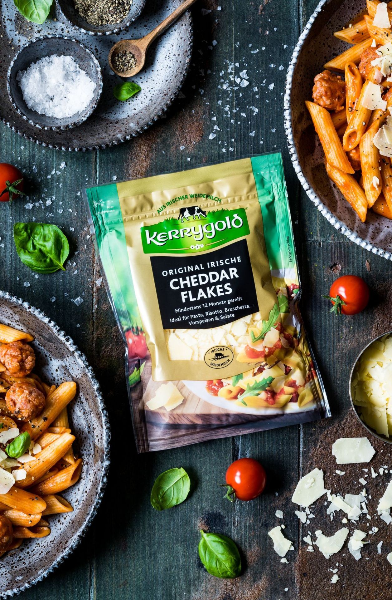 Eine Packung Kerrygold Original Irish Cheddar Flakes auf einem rustikalen Holztisch, umgeben von Nudelgerichten, Kirschtomaten, Basilikumblättern, Käsespänen und kleinen Schüsseln mit Salz und Pfeffer.