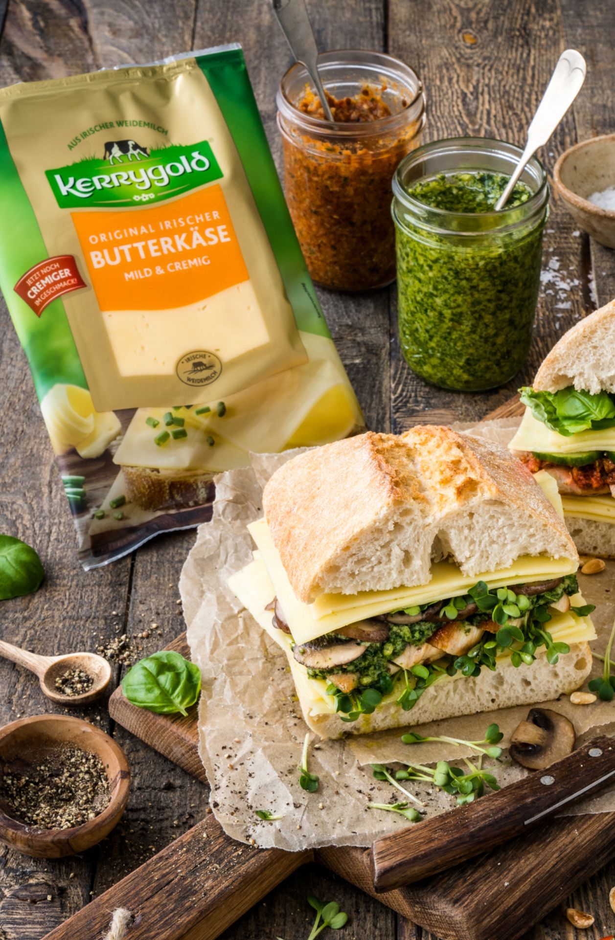 Ein Ciabatta-Sandwich mit Käse, Gemüse und Pilzen liegt auf Backpapier neben Gläsern mit Pesto und Tomatenaufstrich, im Hintergrund eine Packung Kerrygold-Butterkäse. Frische Kräuter und Pfeffer sind darüber verteilt.