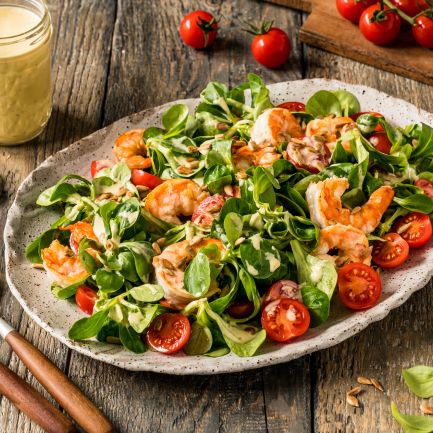 Auf einem rustikalen Holztisch steht ein Teller mit Garnelensalat, Kirschtomaten, Blattgemüse und Dressing, umgeben von einem Glas Dressing, einer kleinen Schüssel mit Samen, Besteck und Strauchtomaten.