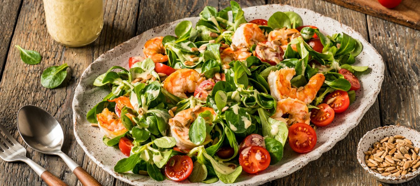 Auf einem rustikalen Holztisch steht ein Teller mit Garnelensalat, Kirschtomaten, Blattgemüse und Dressing, umgeben von einem Glas Dressing, einer kleinen Schüssel mit Samen, Besteck und Strauchtomaten.