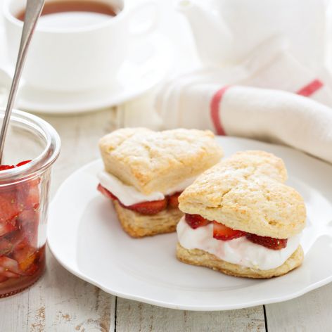 Herzförmige Scones gefüllt mit Sahne und Erdbeeren auf einem weißen Teller erinnern an den Charme eines klassischen Erdbeerkuchens. Neben geschnittenen Beeren und Tee stehen sie auf einem weißen Holztisch mit einer gestreiften Serviette – ein herrlicher deutscher Dessertmoment.