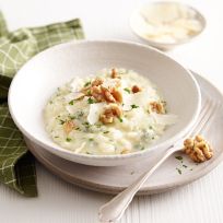 Eine Schale mit cremigem Käse-Risotto, garniert mit Walnüssen und frischer Petersilie. Im Risotto sind Stücke von Käse und Kräutern sichtbar. Rechts neben der Schale liegt eine Gabel, im Hintergrund ist ein grün kariertes Tuch teilweise zu sehen.