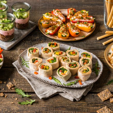 Ein Tisch voller verschiedener Fingerfood-Häppchen, darunter Gemüsewraps mit Frischkäse, gefüllte Mini-Paprika, Grissini, Dips, Gemüsesticks und geschichtete Salatbecher, alles angerichtet auf rustikalen Holzoberflächen.