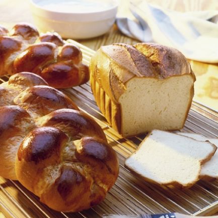 Ein Laib Butterhefezopf und ein Laib Weißbrot, mit einer angeschnittenen Scheibe davor, liegen auf einem Kuchengitter. Daneben liegt ein Backpinsel, im Hintergrund ein helles Tuch und eine Schüssel.