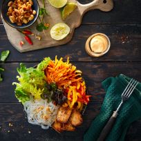 Eine bunte Schüssel vietnamesischer Glasnudelsalat mit mariniertem Tofu, geraspelten Karotten, Salat, Kräutern und Paprika, serviert neben einer kleinen Schüssel mit Erdnüssen, Soße, Limettenspalten und einer Gabel auf einem dunklen rustikalen Tisch.