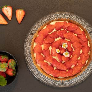 Eine Erdbeer-Tarte mit Sahne steht auf einem Glasteller, dekoriert mit geschnittenen Erdbeeren und weißen essbaren Blüten. Daneben liegen frische Erdbeeren, einige geschnitten, und eine Schale mit ganzen Erdbeeren mit grünen Blättern.