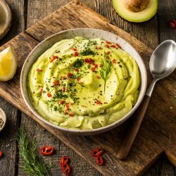 Eine Schüssel mit cremigem Avocadocreme-Dip, garniert mit gehackten roten Chilis, Kräutern und schwarzem Pfeffer, steht auf einem Holzbrett, daneben ein Löffel. Zitronenspalten, Chilischeiben, Kräuter und eine halbierte Avocado sind daneben angeordnet.