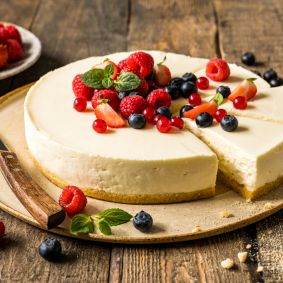 Ein ungebackener Käsekuchen, von dem ein Stück entfernt wurde, garniert mit Erdbeeren, Himbeeren, Blaubeeren, roten Johannisbeeren und Minzblättern, liegt mit einem Messer auf einem Teller. Frische Beeren liegen verstreut auf dem rustikalen Holztisch daneben.