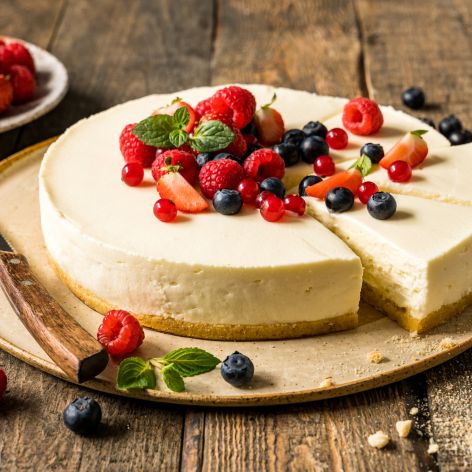 Ein ungebackener Käsekuchen, von dem ein Stück entfernt wurde, garniert mit Erdbeeren, Himbeeren, Blaubeeren, roten Johannisbeeren und Minzblättern, liegt mit einem Messer auf einem Teller. Frische Beeren liegen verstreut auf dem rustikalen Holztisch daneben.
